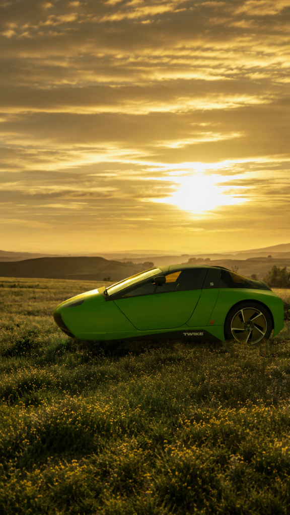 A green TWIKE 5 in a field of flowers against a hilly landscape, bathed in the golden light of the setting sun.